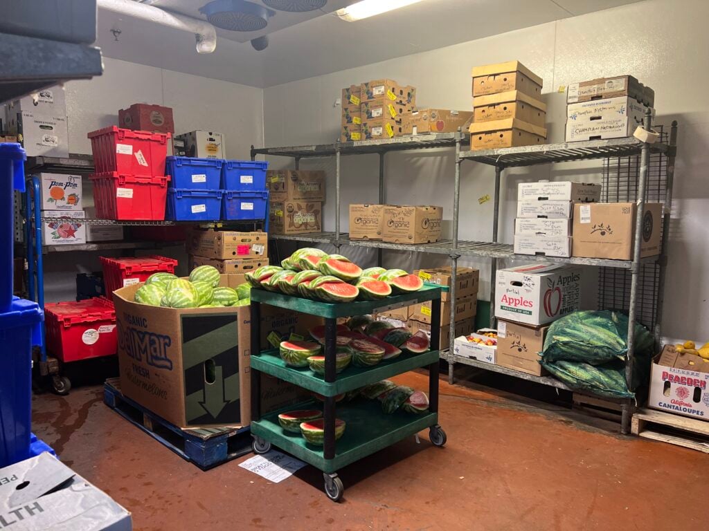 A photo of the walk-in refrigerator in the produce department. A cart with watermelon halves and quarters is in the center, and cardboard boxes filled with vegetables and fruits line the walls, stacked on metal shelves.