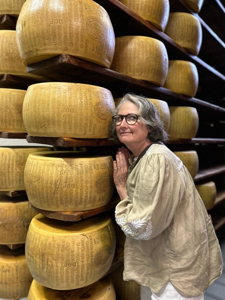 Beth Chevalier manager of the cheese department posing affectionately in a cheese cave amongst many wheels of parmesan aging on wooden shelves in Italy